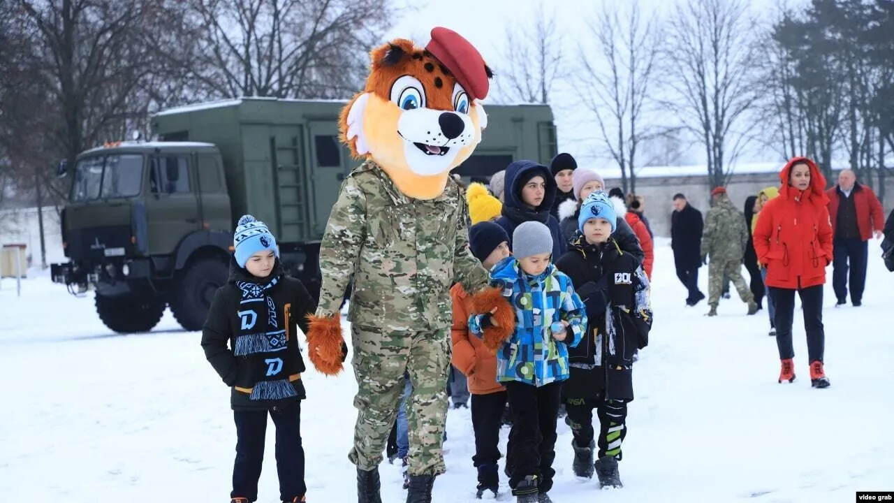 Una fila de niños ucranianos camina tras una persona con camuflaje y un disfraz de tigre-mascota, con un camión militar al fondo — una actividad «militar-patriótica».